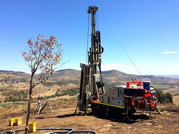 A track-mounted drill rig set up on a rural hillside block with hills behind it